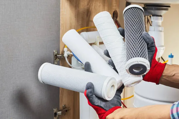 Replacement filter cartridges held in gloved hands, including a carbon block cartridge, in front of an under-sink filtration system.